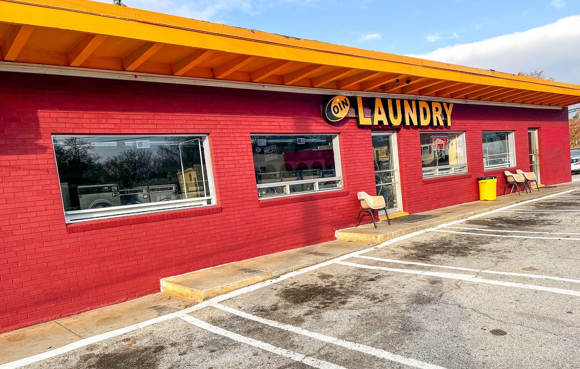 Exterior of a red brick coin laundry building with yellow trim, several windows, and chairs outside.